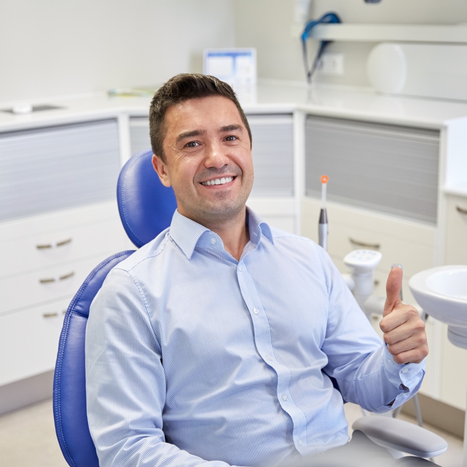 people, medicine, stomatology and health care concept - happy male patient sitting on dental chair and showing thumbs up at clinic office