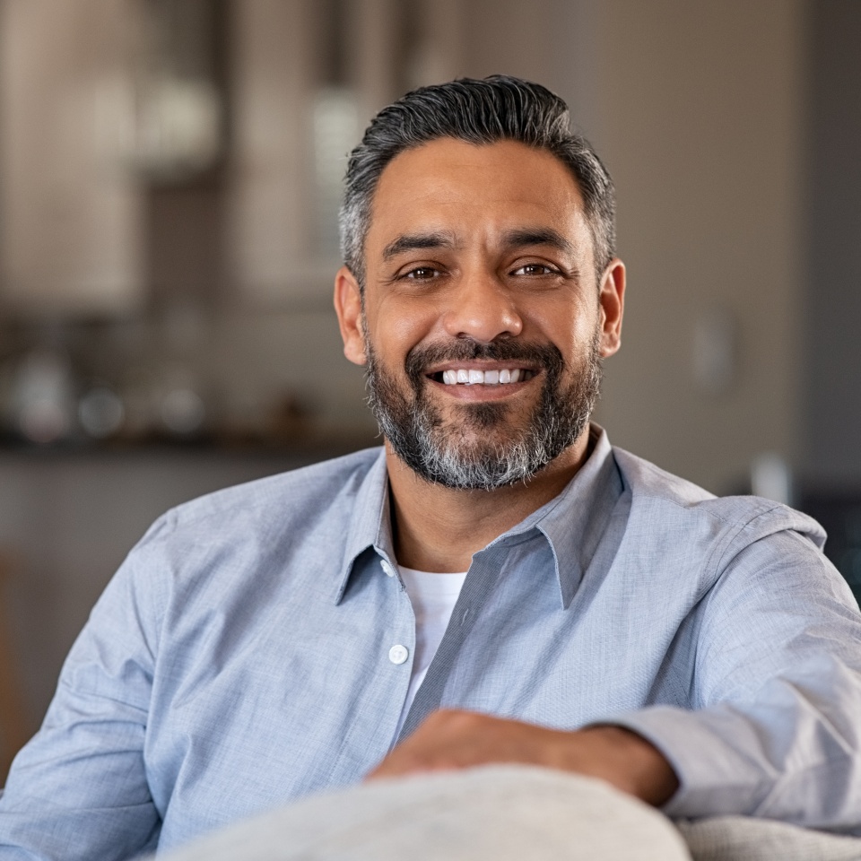 Portrait of happy mid adult man sitting on sofa at home. Handsome latin man in casual relaxing on couch and smiling. Cheerful indian guy looking at camera.