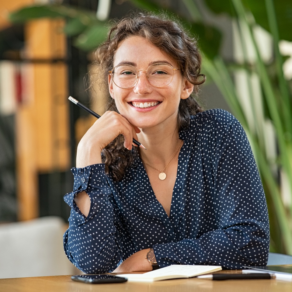 Portrait of smiling woman wearing spectacles while sitting at desk. Business woman taking notes in diary and looking at camera. University girl with eyeglasses sitting on table at library.