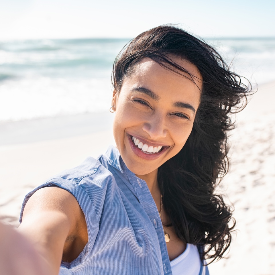 Portrait of smiling young woman taking a selfie at beach during. Cheerful hispanic woman enjoying at beach during holiday. Happy girl taking photo over exotic tropical beach looking at camera.