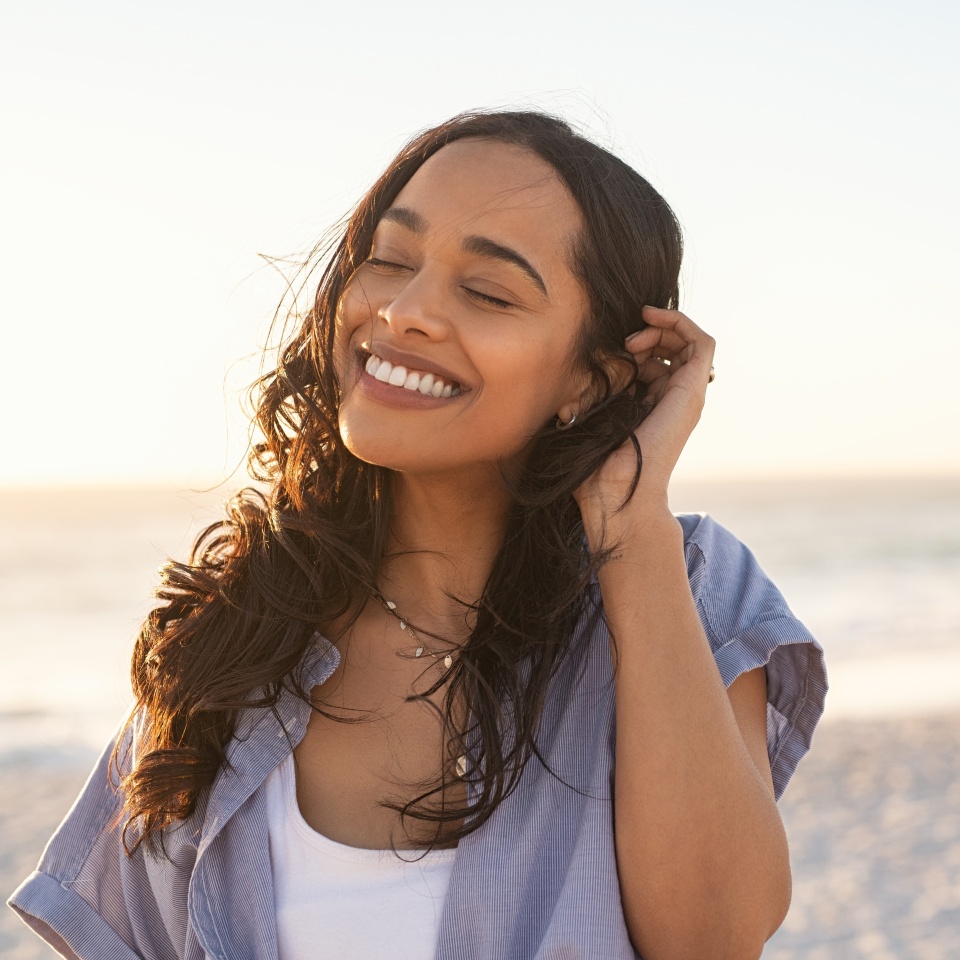 Smiling latin hispanic woman relaxing on beach with closed eyes at sunset. Beautiful mixed race woman enjoying wind fluttering hair. Charming young woman breathing fresh air at summer beach.