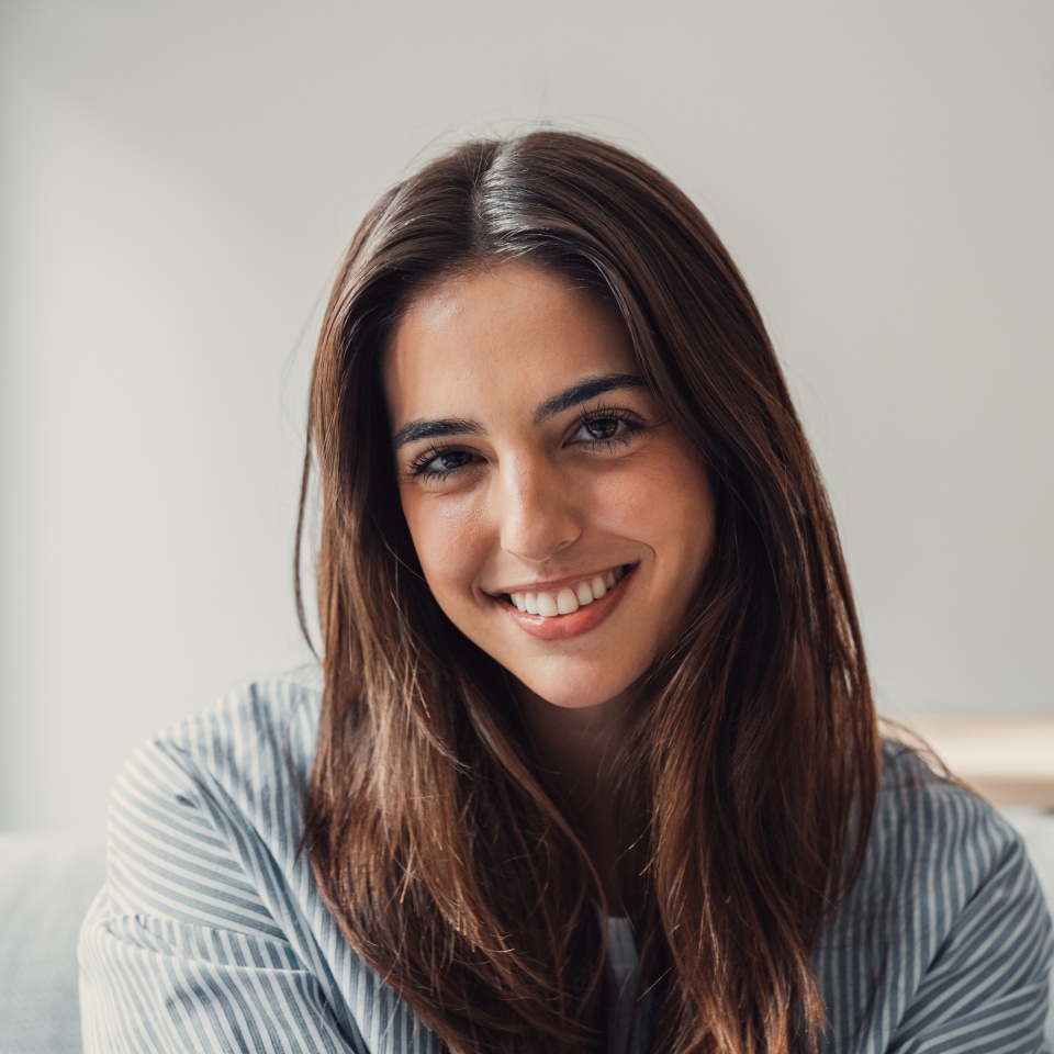 Happy attractive young adult woman housewife sitting on sofa looking at camera in modern cozy home, smiling single 20s lady beautiful face relaxing on couch alone enjoy wellbeing posing for portrait