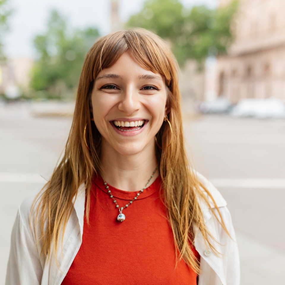 Happy young caucasian girl smiling at camera standing at city street. Outside portrait of joyful beautiful italian woman over urban background.