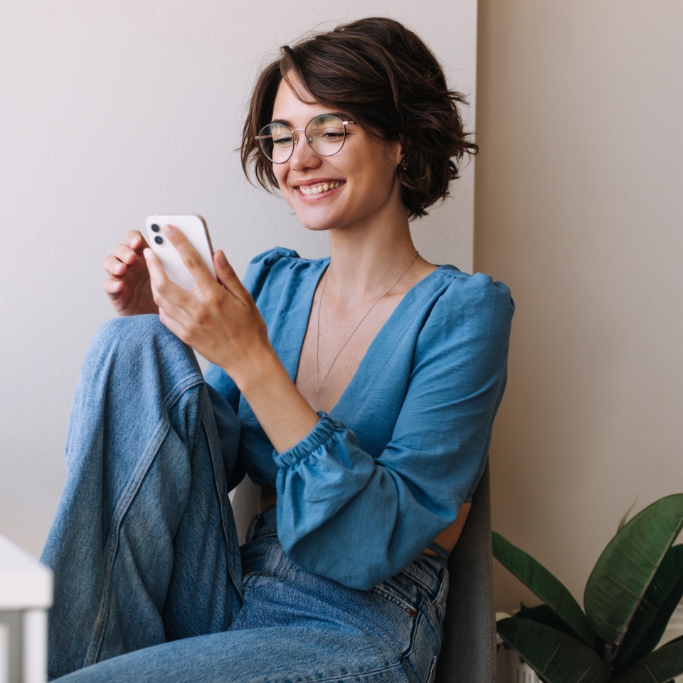 Charming woman holding phone at home . Caucasian smiling brunette woman looking and chatting on smartphone sitting on the floor. Concept of lifestyle, use technology