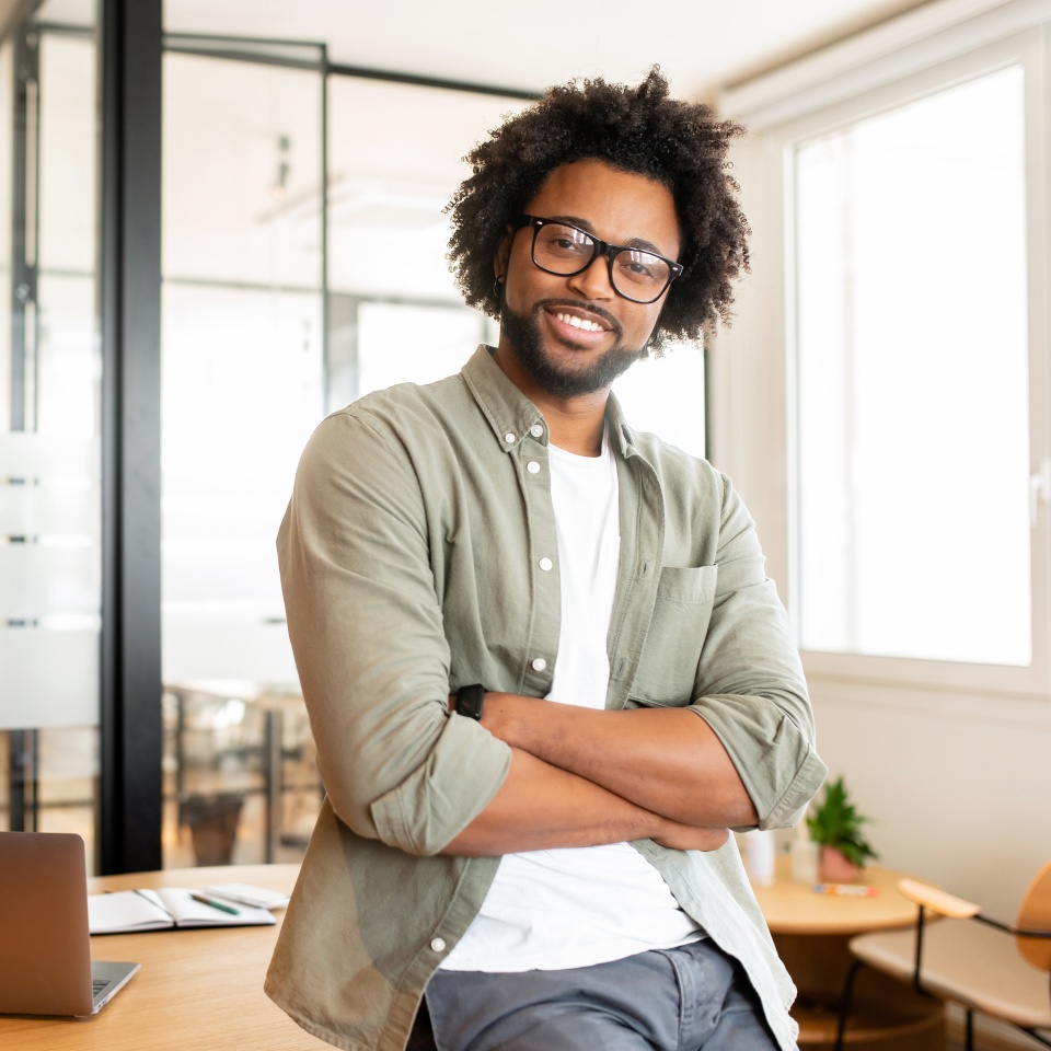 Ambitious highly-skilled handsome curly african-american male employee in glasses and casual wear leaned on office desk and looking at the camera with smile, businessman stands with arms crossed