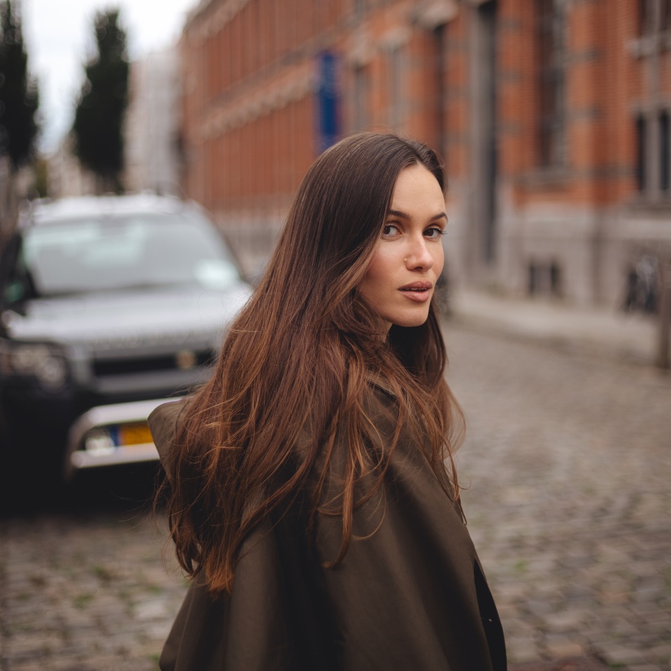 Calm sensual smiling long hair brunette woman cross road, while walking outdoor in old town. Thoughtful woman wear green trench coat. Fashion style outfit.